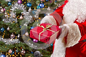 Santa Claus hands holds red gift box opposite christmas tree.