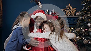 Santa claus in hat sitting on the red chair while five kids unexpectedly running and hugging him.