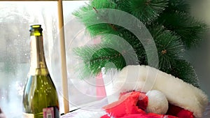 Santa Claus hat on white sill against the window and artificial spruce branches. Red hat, opened bottle of sparkling