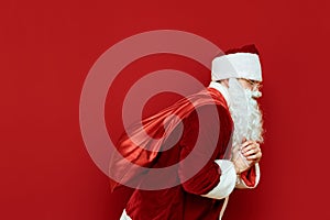 Santa Claus with a heavy bag of gifts on his back goes on a red background, looking away. Santa and blank space for text. Isolated