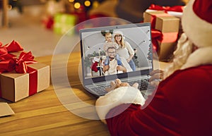 Santa Claus in his workshop video calling young family using modern laptop computer