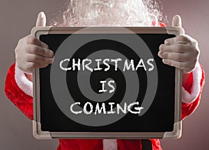 Santa Claus holding a black chalk board written with CHRISTMAS IS COMING