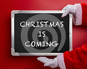 Santa Claus holding a black chalk board written with CHRISTMAS IS COMING on red