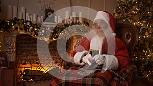 Santa Claus holds a knitted sock in his hands near fireplace. Santa is resting after a hard day, knitting a woolen sock