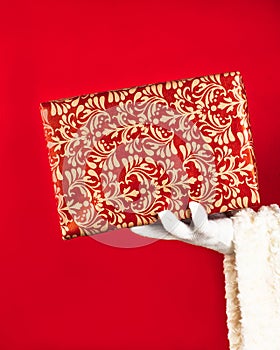 Santa Claus holds a paper box with gift in his hand on a red background with copy space. Giving a gift concept