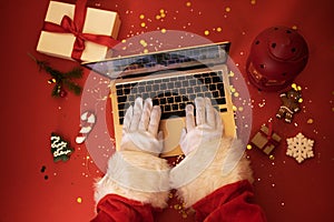 Santa Claus with laptop. Hands in white gloves typing on an notebook keyboard. Gift boxes, christmas cookies, lantern and fir