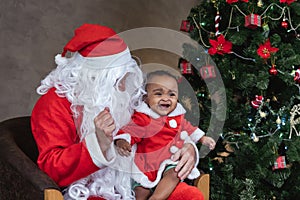 Santa Claus is lifting happy little toddler baby girl up and laughing cheerfully with fully decorated christmas tree on the back