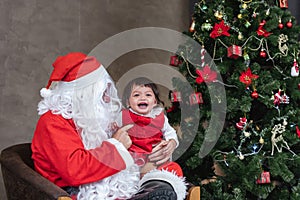 Santa Claus is lifting happy little toddler baby girl up and laughing cheerfully with fully decorated christmas tree on the back