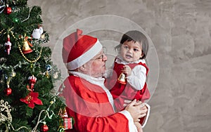 Santa Claus is lifting happy toddler baby girl up and laughing cheerfully while helping to decorate christmas tree on the back for