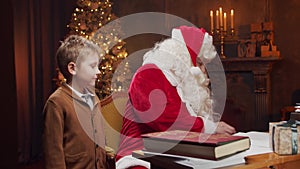 Santa Claus and little boy. Cheerful Santa is working while sitting at the table. Fireplace and Christmas Tree in the