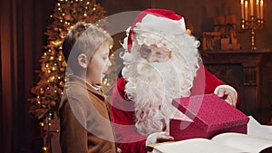 Santa Claus and little boy. Cheerful Santa is working while sitting at the table. Fireplace and Christmas Tree in the