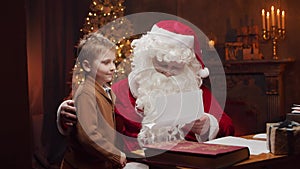 Santa Claus and little boy. Cheerful Santa is working while sitting at the table. Fireplace and Christmas Tree in the