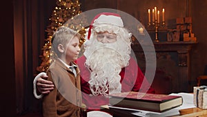 Santa Claus and little boy. Cheerful Santa is working while sitting at the table. Fireplace and Christmas Tree in the