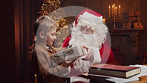 Santa Claus and little boy. Cheerful Santa is working while sitting at the table. Fireplace and Christmas Tree in the