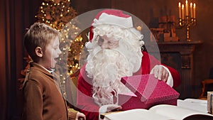 Santa Claus and little boy. Cheerful Santa is working while sitting at the table. Fireplace and Christmas Tree in the