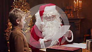 Santa Claus and little boy. Cheerful Santa is working while sitting at the table. Fireplace and Christmas Tree in the