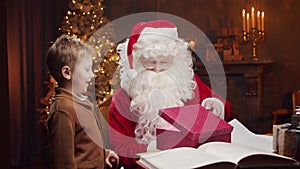 Santa Claus and little boy. Cheerful Santa is working while sitting at the table. Fireplace and Christmas Tree in the