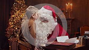 Santa Claus and little boy. Cheerful Santa is working while sitting at the table. Fireplace and Christmas Tree in the