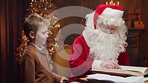 Santa Claus and little boy. Cheerful Santa is working while sitting at the table. Fireplace and Christmas Tree in the