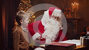 Santa Claus and little boy. Cheerful Santa is working while sitting at the table. Fireplace and Christmas Tree in the