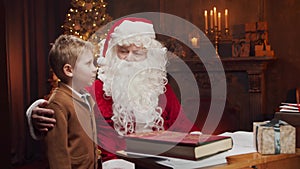 Santa Claus and little boy. Cheerful Santa is working while sitting at the table. Fireplace and Christmas Tree in the