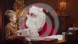 Santa Claus and little boy. Cheerful Santa is working while sitting at the table. Fireplace and Christmas Tree in the