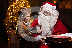 Santa Claus and little boy. Cheerful Santa is working while sitting at the table. Fireplace and Christmas Tree in the