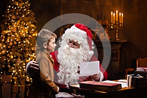 Santa Claus and little boy. Cheerful Santa is working while sitting at the table. Fireplace and Christmas Tree in the