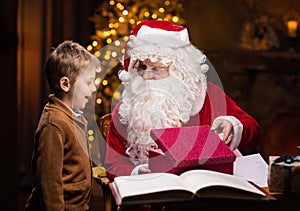 Santa Claus and little boy. Cheerful Santa is working while sitting at the table. Fireplace and Christmas Tree in the