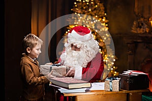 Santa Claus and little boy. Cheerful Santa is working while sitting at the table. Fireplace and Christmas Tree in the