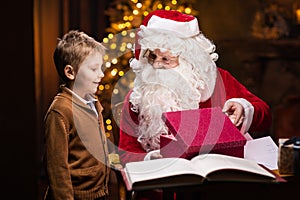 Santa Claus and little boy. Cheerful Santa is working while sitting at the table. Fireplace and Christmas Tree in the