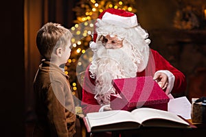 Santa Claus and little boy. Cheerful Santa is working while sitting at the table. Fireplace and Christmas Tree in the