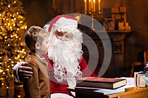 Santa Claus and little boy. Cheerful Santa is working while sitting at the table. Fireplace and Christmas Tree in the