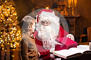 Santa Claus and little boy. Cheerful Santa is working while sitting at the table. Fireplace and Christmas Tree in the