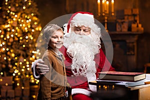 Santa Claus and little boy. Cheerful Santa is working while sitting at the table. Fireplace and Christmas Tree in the