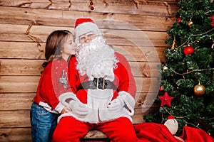 Santa Claus and Little girl. Girl telling wish in Santa Claus`s ear in front of Christmas Tree