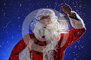 Santa Claus with a long white beard holds candle holder with burning candle against a snowing blue sky. Christmas