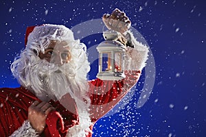 Santa Claus with a long white beard holds a lamp with a candle against a blue sky. It is snowing. Christmas