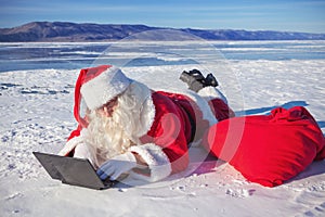 Santa Claus lying on the snow, looking at laptop n