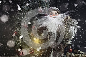Santa Claus Outdoors Beside Christmas Tree in Snowfall Carrying Gifts to Children