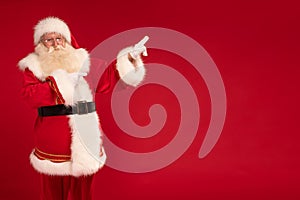 Santa Claus pointing on empty red studio background. Merry Christmas and Happy New Year! A lot of copy space. Xmas sale, discount