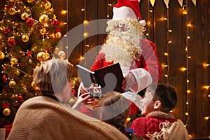 Santa Claus reading book for family. Mother and children sitting indoor near decorated xmas tree with lights - Merry Christmas and