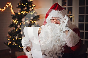 Santa Claus reading from a long list, in a cozy room