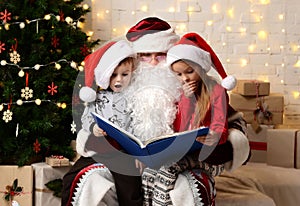Santa Claus reading magic book to happy little cute children boy and girl kids near Christmas tree