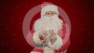 Santa claus reads and sends text messages from his cell phone on red background with snow