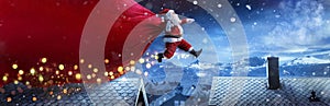 Santa Claus With Red Big Bag Jumping On Roofs In Snowy Winter Landscape