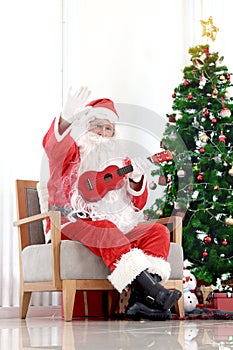 Santa Claus in red costume playing Ukulele and singing Christmas songs front of decorative Christmas tree in living room. Happy