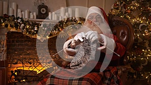 Santa Claus showing a knitted sock in his hands near fireplace. Santa is resting after a hard day, knitting a woolen