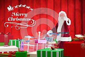 Santa claus showing thumbs up while holding shopping cart full of gifts