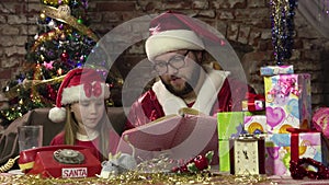 Santa Claus sits on armchair and reads book with fairy tales for enthusiastic little girl